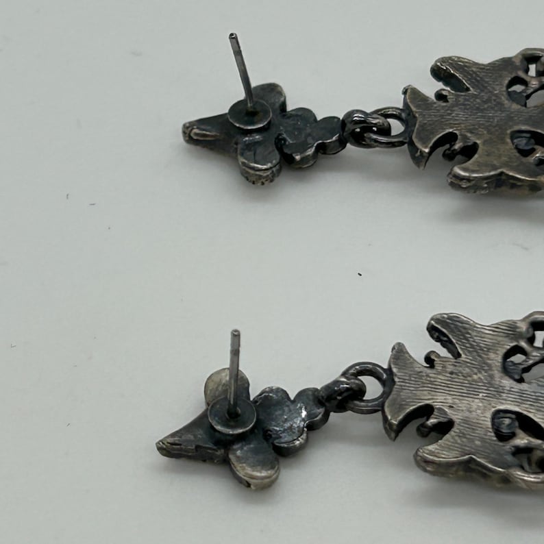 Puede incluir: Primer plano de dos pendientes colgantes plateados. Cada pendiente presenta un perno floral, conectado a una gota ornamentada. Los pendientes tienen una est&eacute;tica vintage o antigua.