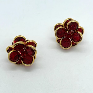 May include: A pair of stud earrings featuring a flower-shaped design. Each earring is crafted with deep red circular crystals set in a polished gold-tone metal frame, arranged in a layered petal pattern that creates a dimensional, rounded appearance.