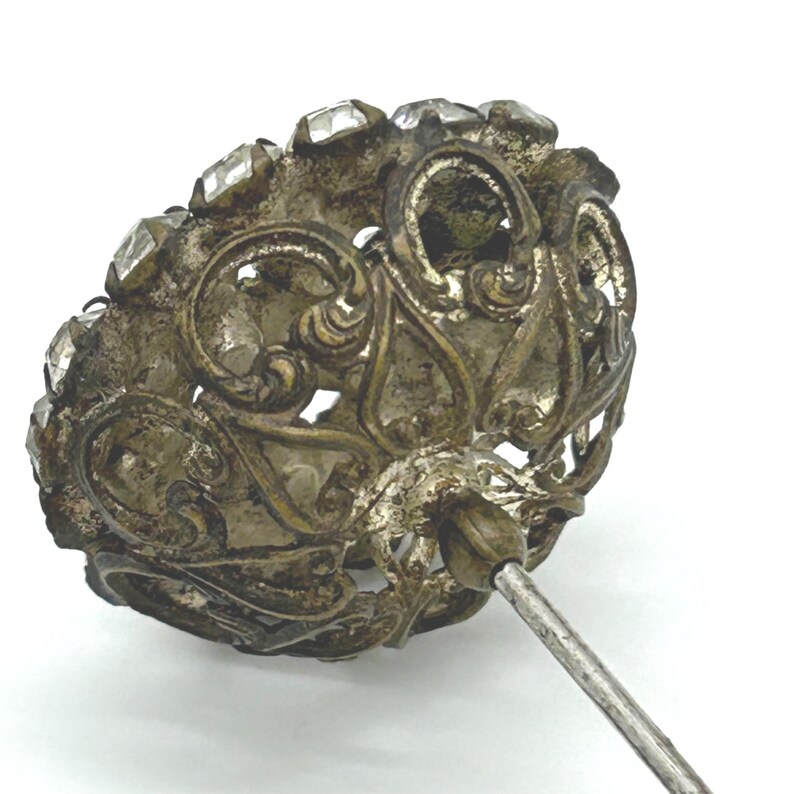 May include: An antique silver-toned hat pin with a decorative head. The head features clear, faceted stones set around an ornate, openwork design. The pin has a long, slender, silver-colored shaft.
