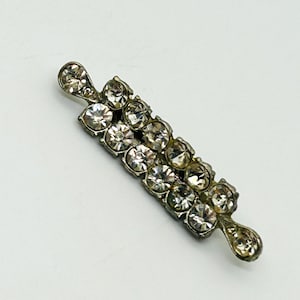 May include: A silver-toned metal bar brooch featuring two parallel rows of clear, round, faceted rhinestones. The brooch has a slender, elongated design with a single, smaller, teardrop-shaped rhinestone set at each of the two pointed ends.