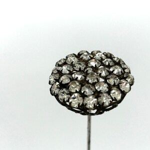 May include: A vintage hat pin with a cluster of clear, faceted rhinestones. The round head is set in a dark metal filigree, mounted on a slender, silver-toned pin. The pin is set against a white background.