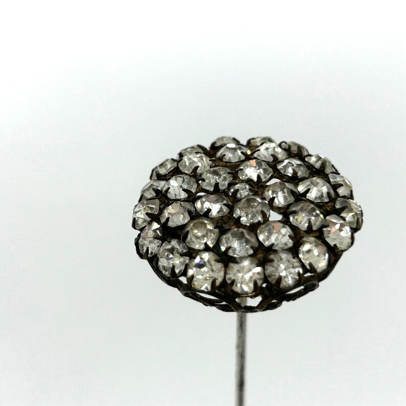 May include: A vintage hat pin with a cluster of clear, faceted rhinestones. The round head is set in a dark metal filigree, mounted on a slender, silver-toned pin. The pin is set against a white background.