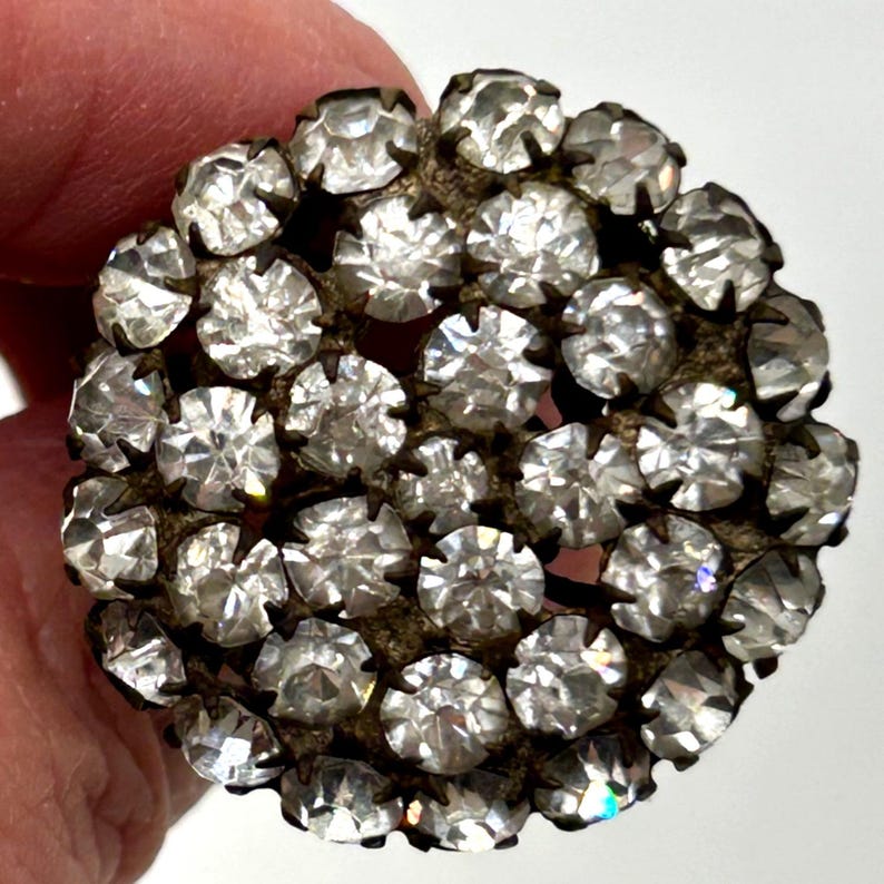 May include: Vintage round brooch with a cluster of clear, faceted rhinestones. The rhinestones are set in a tarnished, aged metal base. The brooch has a classic, elegant design, perfect for adding sparkle to any outfit.