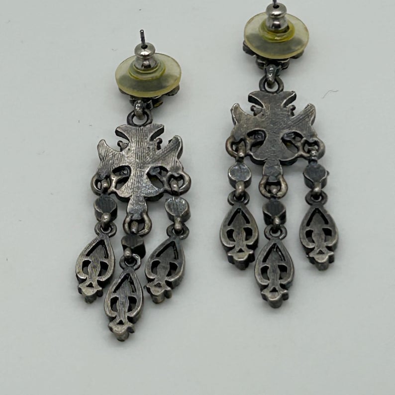 Puede incluir: Un par de pendientes colgantes plateados. Cada pendiente presenta una parte superior decorativa en forma de cruz con colgantes en forma de l&aacute;grima. Los pendientes tienen cierre de tuerca y est&aacute;n sobre un fondo blanco.