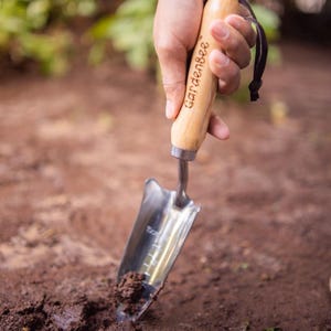 May include: A hand holds a small gardening trowel with a wooden handle. The handle is engraved with the words "gardenbee". The trowel is digging into the brown soil.
