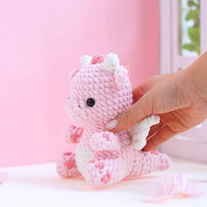 May include: A handmade pink crochet dragon with white accents on its wings, horns, and belly. The plush toy has black safety eyes and is sitting on a light pink surface. The dragon is a cute, whimsical design.