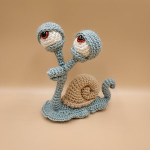 May include: A whimsical, handmade crochet snail. The snail features large, googly eyes with red pupils, a tan shell, and a light blue body. The snail is made of soft yarn and is a unique decorative item.