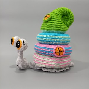May include: A whimsical, handmade crochet snail toy. The snail features a stack of colorful, striped rings in shades of pink, blue, and green, topped with a green hat. The snail's body is gray, with large, expressive eyes.