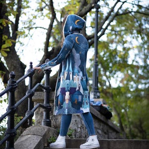 May include: A woman wearing a blue hooded cardigan with a whimsical city scene print. The cardigan has a hood with a white lining and a white trim around the edges. The woman is walking down a set of stone steps.