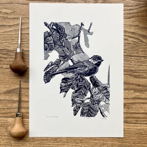 Op de afbeelding: Een zwart-wit linoleumsnede van een vogel op een tak met bladeren. De prent is gesigneerd en genummerd. Twee gereedschappen met houten handvatten zijn aanwezig, wat duidt op de druktechniek. Het kunstwerk staat op wit papier.
