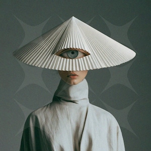 Puede incluir: Un sombrero cónico blanco plisado de ala ancha con un gráfico realista de un ojo humano centrado en la parte delantera. El sombrero se lleva sobre una prenda de color gris claro con cuello alto que cubre el cuello y los hombros.