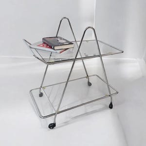 Könnte beinhalten: Ein verchromter Servierwagen aus Metall mit zwei Glasböden und vier schwarzen Rollen. Ein Buch liegt auf der oberen Ablage.