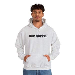 May include: A light gray hooded sweatshirt with the text "NAP QUEEN" in black block letters. The sweatshirt has a front pocket and a drawstring hood. The person is wearing blue jeans.