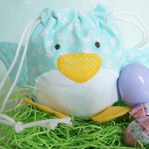 以下が含まれることがあります： 黄色いクチバシと白い毛皮を持つ、青と白の布製のイースターバニーバッグ。バッグは、パステルパープルとピンクのイースターエッグとチョコレートエッグでいっぱいです。