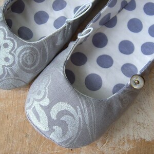 May include: A pair of gray fabric ballet flats with a silver floral pattern on the outside and a white background with gray polka dots on the inside. One flat has a button on the side.
