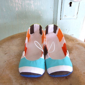 May include: A pair of white, brown, orange, and turquoise striped fabric flats. The shoes have a white elastic strap across the top.