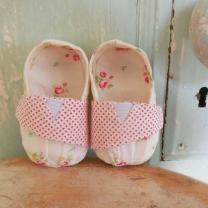 Pode incluir: Um par de sapatos de bebé brancos com acabamento em bolinhas cor-de-rosa e um padrão floral. Os sapatos são feitos de tecido e têm uma sensação macia e confortável.
