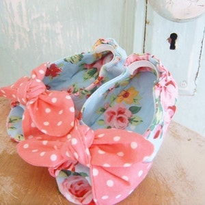 Pode incluir: Um par de sapatos de bebé com laços de bolinhas rosa e brancas. Os sapatos são feitos de um tecido azul claro com estampa floral.