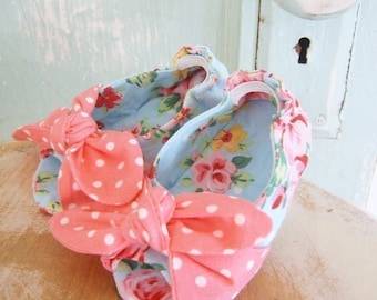 Op de afbeelding: Een paar babyschoenen met roze en witte stippenboogjes. De schoenen zijn gemaakt van lichtblauwe stof met een bloemenprint.
