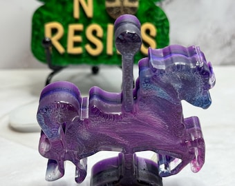 May include: A purple and blue resin carousel horse figurine. The horse has a swirling, marbled effect with shades of purple and blue. The base is black. The background features a green sign with the words "DOUBLE N RESINS" in yellow.