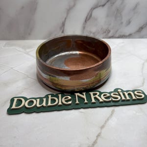May include: A small, round bowl with a copper and silver marbled design. The bowl has a green, yellow, and white marbled rim. The bowl is sitting on a white marble surface. The text "Double N Resins" is visible in the foreground.