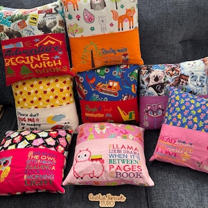 May include: A collection of colorful, embroidered reading pillows with various designs. Pillows feature text like "Adventure Begins with Books" and "Llama Likes Drama." Designs include foxes, owls, cats, and construction vehicles. The pillows are on a gray surface.