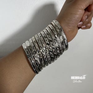 May include: A set of silver bangles, each adorned with detailed etched patterns. The bangles are stacked on a wrist, displaying a range of designs, including geometric and floral elements. The jewellery is presented against a white backdrop.