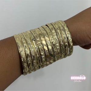 May include: A collection of gold bangles with intricate engraved patterns. The bangles are stacked on a wrist, showcasing their detailed designs and shiny finish. The image is set against a plain background.
