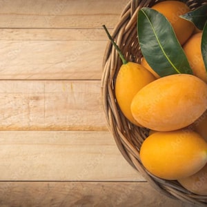 May include: A woven basket filled with ripe, oval-shaped yellow fruits, possibly Marian plums, with green leaves. The basket sits on a light brown wooden surface, creating a natural, rustic aesthetic.