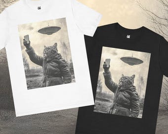 May include: Two t-shirts with a black and white image of a cat taking a selfie with a flying saucer in the background. The cat is wearing a coat and holding a smartphone. The t-shirts are one white and one black.