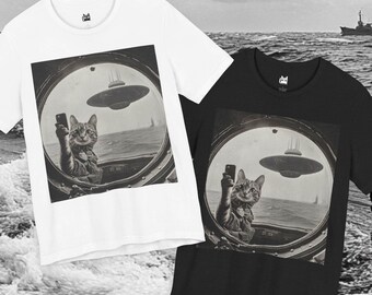 May include: White and black t-shirts featuring a vintage-style image of a cat taking a selfie with a UFO in the background. The cat is inside a circular window, with the ocean and a ship visible.