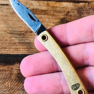 Op de afbeelding: Een klein vouwmes met een messing handvat en een stalen lemmet met patroon. Het lemmet is open en onthult een gedetailleerd ontwerp. Het handvat heeft een gebogen vorm en twee ronde gaten. Het mes wordt in een hand gehouden.