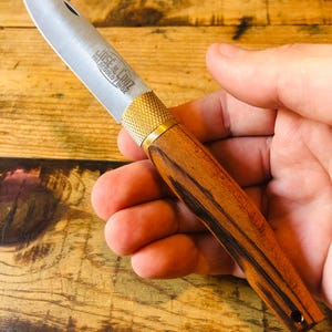 Peut inclure: Un couteau de poche pliant avec une lame en acier inoxydable argenté portant l'inscription gravée JOSE DA CRUZ PALACUOLO INOX. Le manche est en bois brun avec un motif de grain sombre et une virole en métal doré texturé.