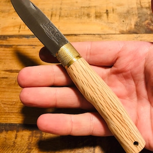 Peut inclure: Un couteau de poche pliant avec une lame en acier argenté portant l'inscription gravée Jose da Cruz, une mitre en laiton texturé et un manche en bois clair avec un grain visible et un petit trou circulaire à la base.