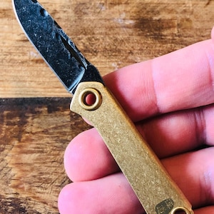 May include: A small folding knife with a dark, textured blade and a brass handle. The blade has a unique pattern and a partially serrated edge. The handle features two circular holes and a small, stamped design. The knife is compact and appears to be a pocket knife.