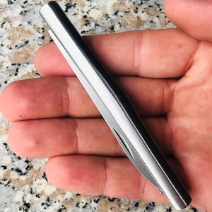 May include: A small, silver pocket knife with a sleek, minimalist design. The knife is closed, showing a smooth, metallic surface. The blade is partially visible. The knife is held in an open hand, showcasing its size and form. The knife is approximately 10 cm long.