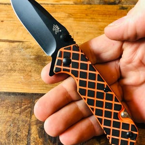 Puede incluir: Una navaja plegable con una hoja negra mate afilada que presenta un pequeño logotipo y un mango texturizado. El mango tiene una rejilla de patrón de diamantes de color naranja brillante sobre una base negra, fijada con tornillos negros.