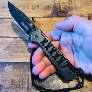 May include: A black folding knife with a silver handle and a black cord. The handle has a decorative black crisscross pattern. The blade is open and angled towards the top left. The knife is held in a hand.