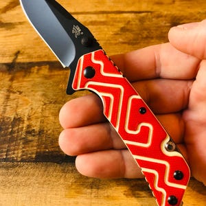 May include: A folding pocket knife with a matte black blade featuring a small logo and a textured handle with a vibrant red and cream-colored geometric zigzag pattern. The handle is secured with black screws and includes a thumb stud for opening.
