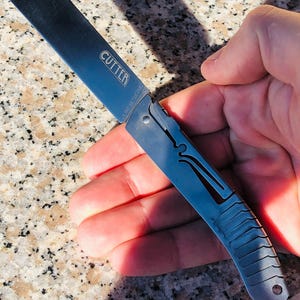 May include: A silver-toned folding utility knife with a flat, rectangular blade featuring the word CUTTER and the text US PAT 10,710,253. The handle has a metallic finish with a textured, grooved grip area and a cutout design along the side frame.
