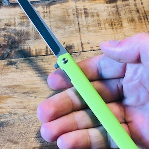 Op de afbeelding: Een neon groen zakmes met een zwart lemmet. Het mes wordt in een hand gehouden, waarbij het handvat en het gedeeltelijk zichtbare lemmet te zien zijn. Het mes heeft een strak, modern design.