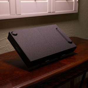 Puede incluir: Un soporte inclinado negro de textura rugosa para equipos electrónicos, con una superficie rectangular que presenta una hendidura circular en la parte superior izquierda y una ranura estrecha y alargada en el lado derecho. La marca AKAI aparece en el lateral.
