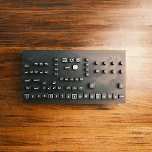 Puede incluir: Un dispositivo negro de producción de música electrónica con numerosos botones, perillas y una pantalla. El dispositivo tiene forma rectangular y está colocado sobre una superficie de madera. Los botones están etiquetados con texto y símbolos.
