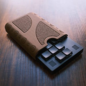 以下が含まれることがあります： 茶色の革製カードホルダー。テクスチャ加工されたグリップと黒いカードインサートが付いています。カードホルダーは、丸みを帯びたエッジと安全な留め具を備えたユニークなデザインです。黒いカードインサートには、いくつかの正方形のボタンがあります。