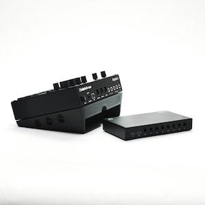 May include: A black Elektron Digitakt II drum computer and sampler mounted on an angled stand, positioned next to a separate black rectangular audio interface unit featuring multiple input ports and two USB-C connections on the front panel.