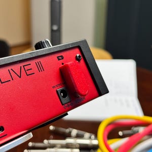 Puede incluir: Primer plano de un dispositivo de audio rojo con el texto "PC LIVE III" en blanco. El dispositivo tiene una parte superior negra con una perilla y una cubierta roja sobre un puerto. Varios cables y conectores son visibles en primer plano.