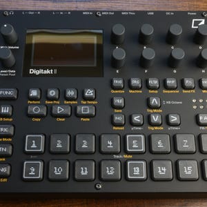 May include: Black Digitakt II electronic music production device with numerous knobs, buttons, and a screen. Features include function buttons, track controls, and a numeric keypad. The device is designed for music production and live performance.
