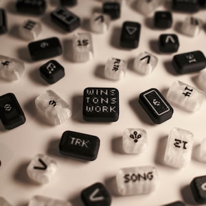 Puede incluir: Vista aérea de fichas de juego rectangulares dispersas. Algunas son negras con texto o símbolos blancos, otras translúcidas con texto negro. Una ficha dice "WINS TONS WORK".