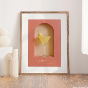 May include: A framed print featuring a Lychee Martini cocktail. The artwork has a peach-colored background with an arch-shaped cutout showcasing the drink. The cocktail is garnished with a yellow fruit. The frame is made of wood.