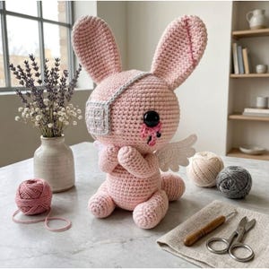 May include: A handmade pink crochet bunny with a gray eye patch and small white wings. The bunny has a black eye and a tear. A ball of pink yarn, a crochet hook, and scissors are also visible.